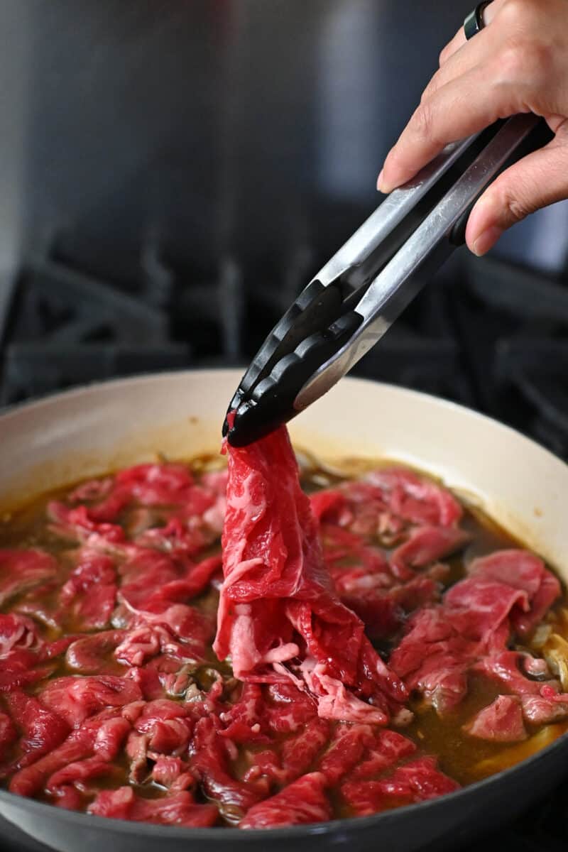 A pair of tongs is adding thinly sliced raw beef to a skillet filled with simmering brown sauce and sliced onions.