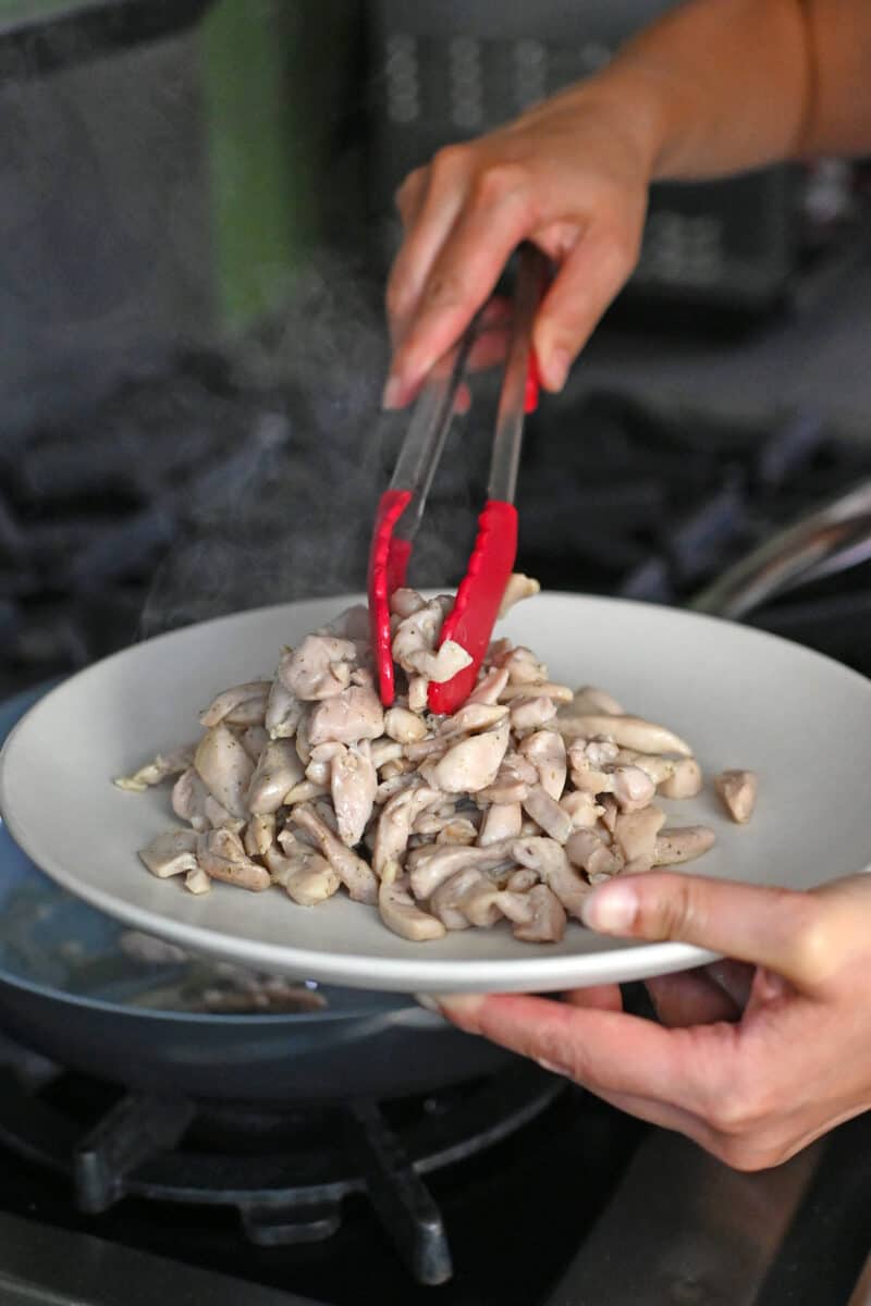 Using a pair of tongs to transfer cooked thinly sliced chicken thighs to a plate for Moo Shu Chicken.