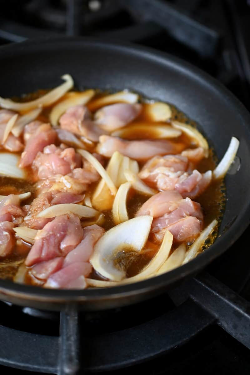 Closeup of a skillet filled with onion slices and raw chicken pieces in a brown sauce.