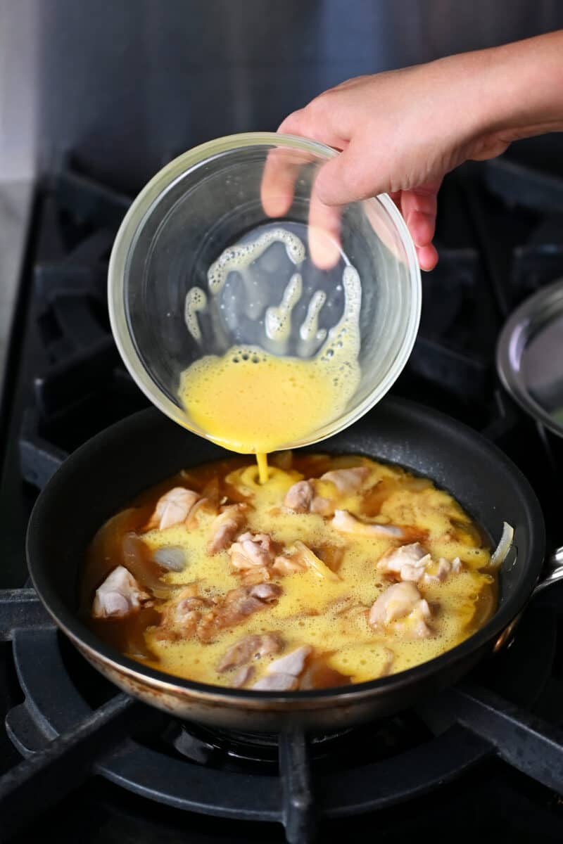 Adding a whisked raw egg to a skillet filled with cooked chicken bites, sliced onion, and a paleo brown sauce.