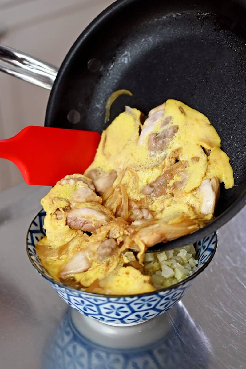 A red silicone spatula is transferring cooked chicken, egg, and sauce to a blue and white bowl filled with cauliflower rice. 