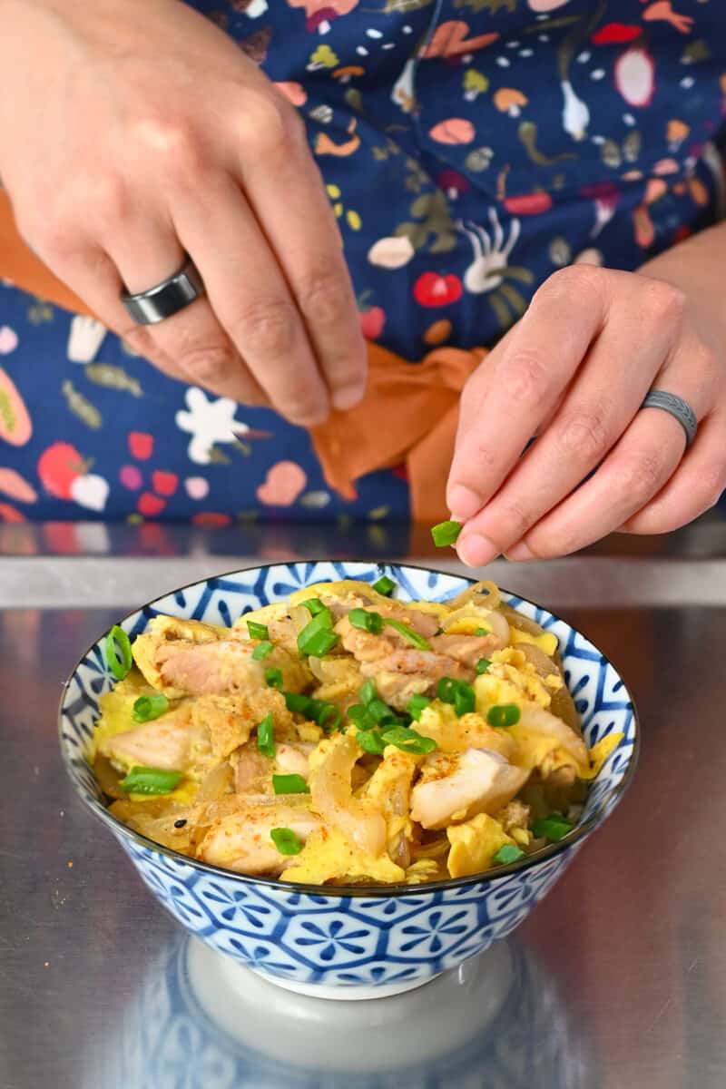 Sprinkling sliced green onions or scallions onto a bow l filled with gluten free oyakodon, Japanese chicken and egg bowl.