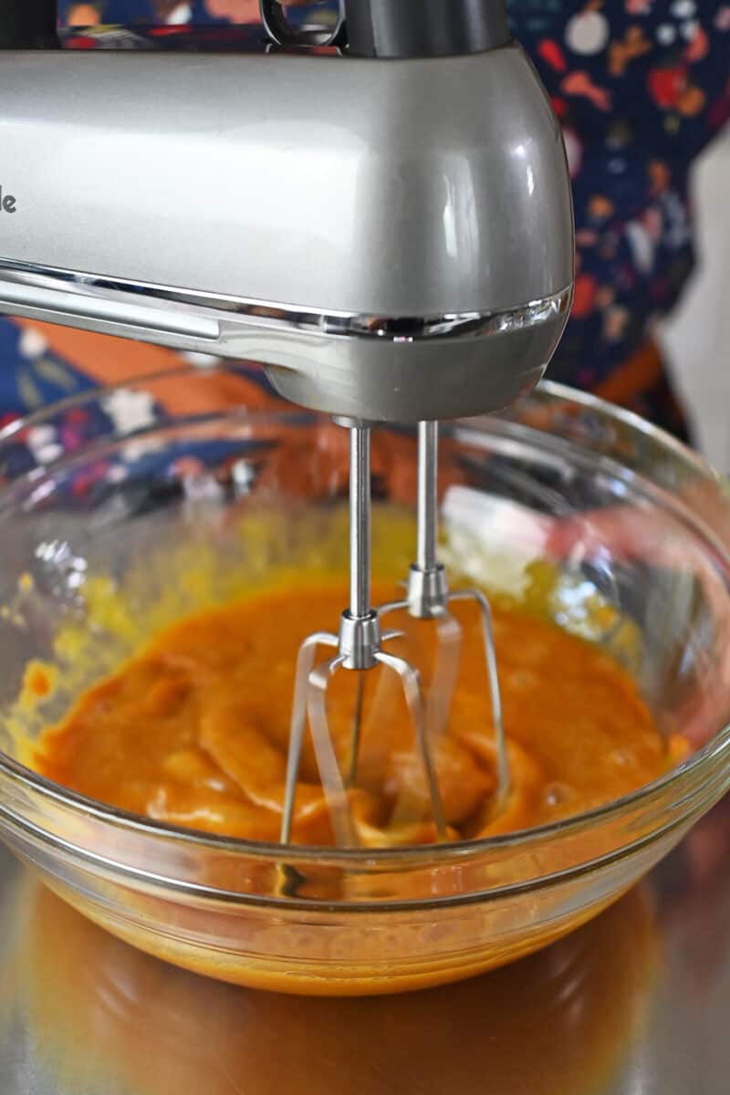 Using a hand mixer to combine the wet ingredients for paleo pumpkin bars in a clear glass bowl.