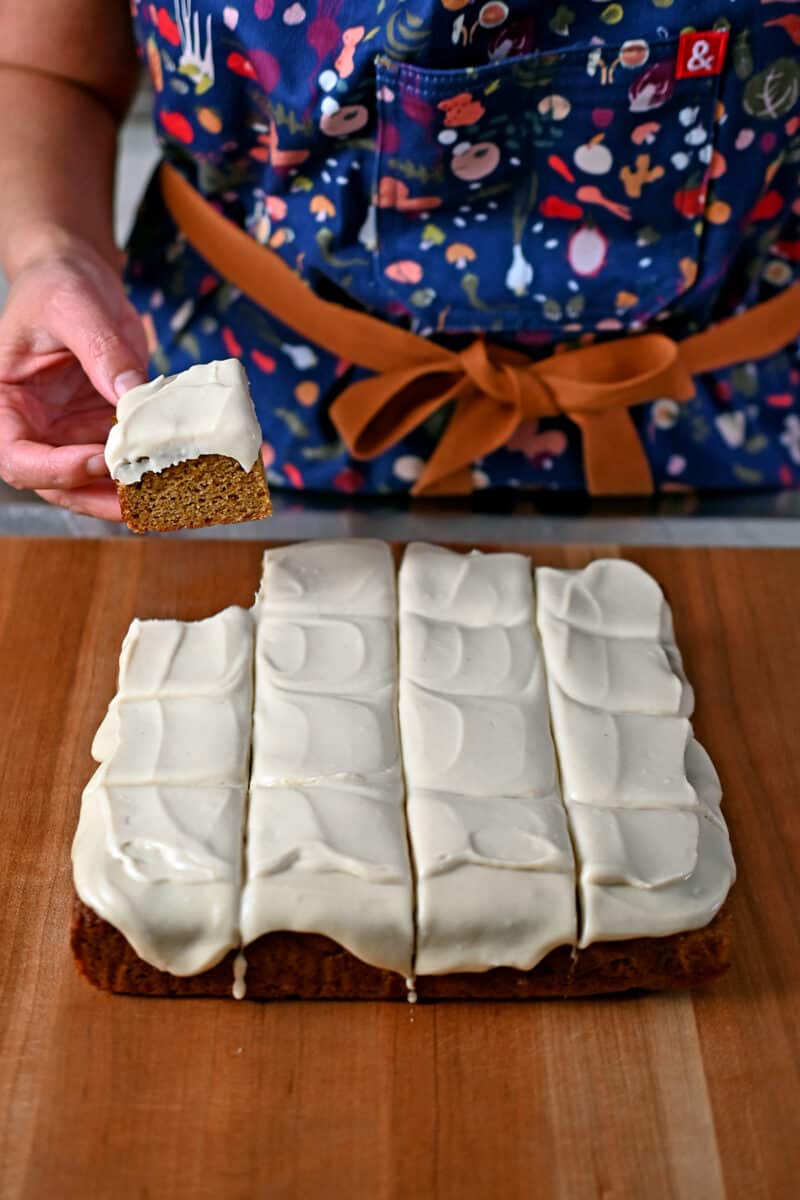 A person in an apron lifting up a pumpkin bar with vegan cream cheese frosting on top.
