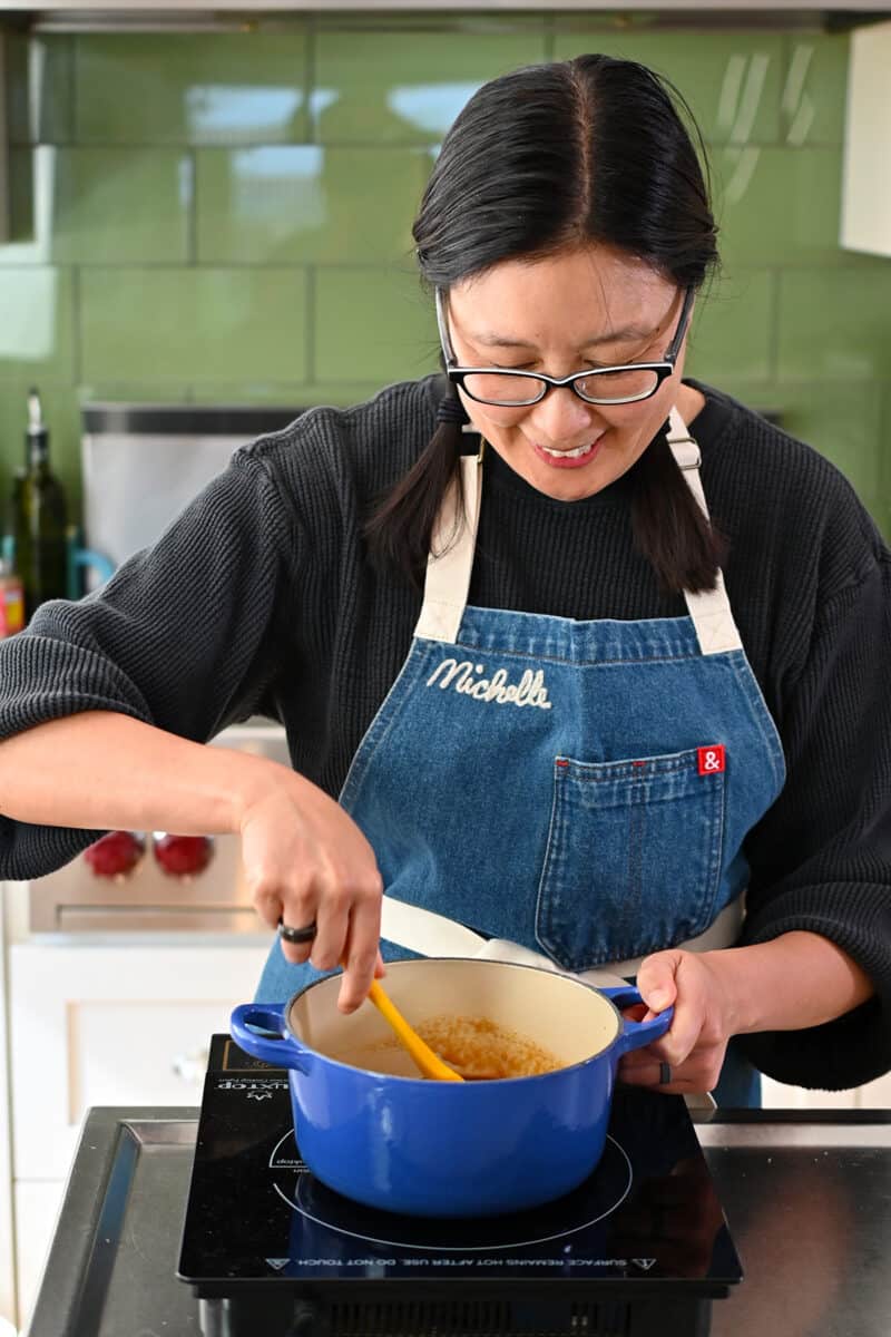 A smiling Asian woman is stirring homemade Tonkatsu Sauce simmering in a blue saucepan.