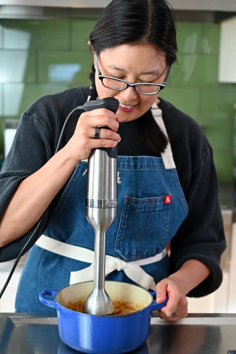 A smiling Asian woman is using an immersion blender to puree homemade Tonkatsu Sauce in a small blue saucepan.