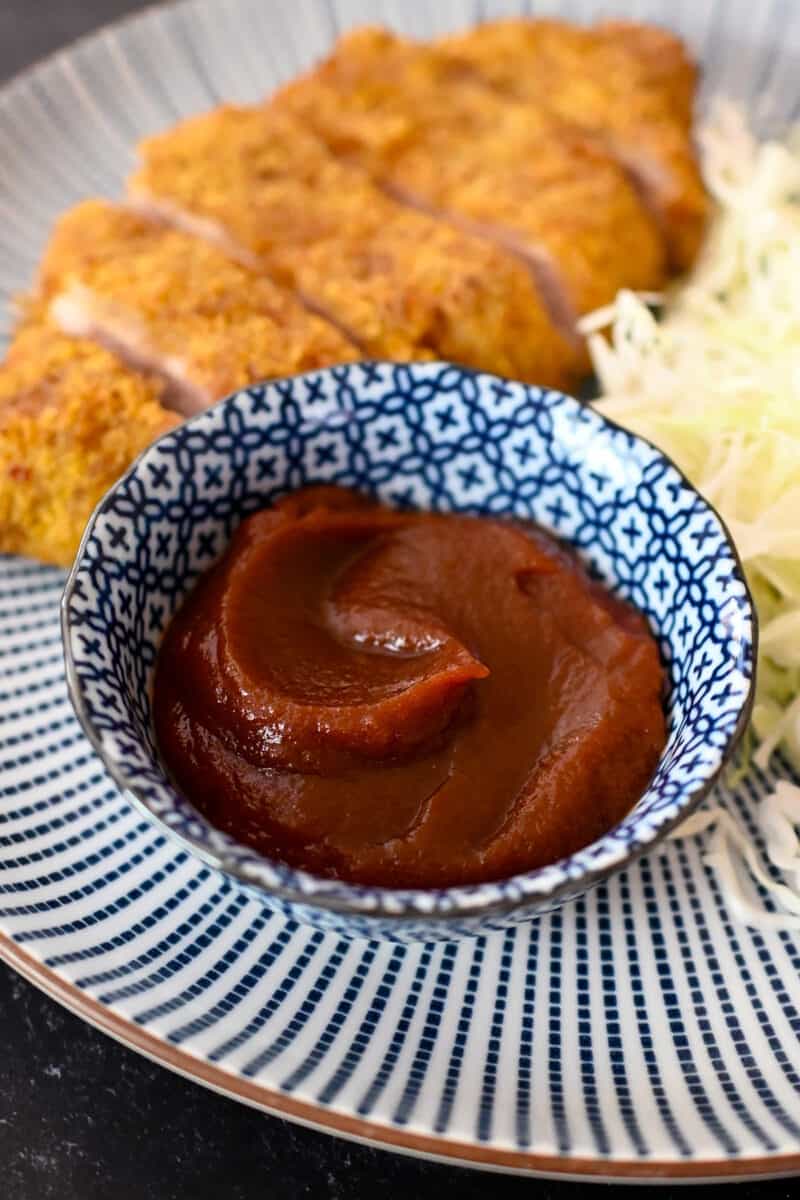 A closeup shot of a small blue bowl filled with homemade Tonkatsu Sauce next to a fried Japanese pork cutlet and shaved cabbage.