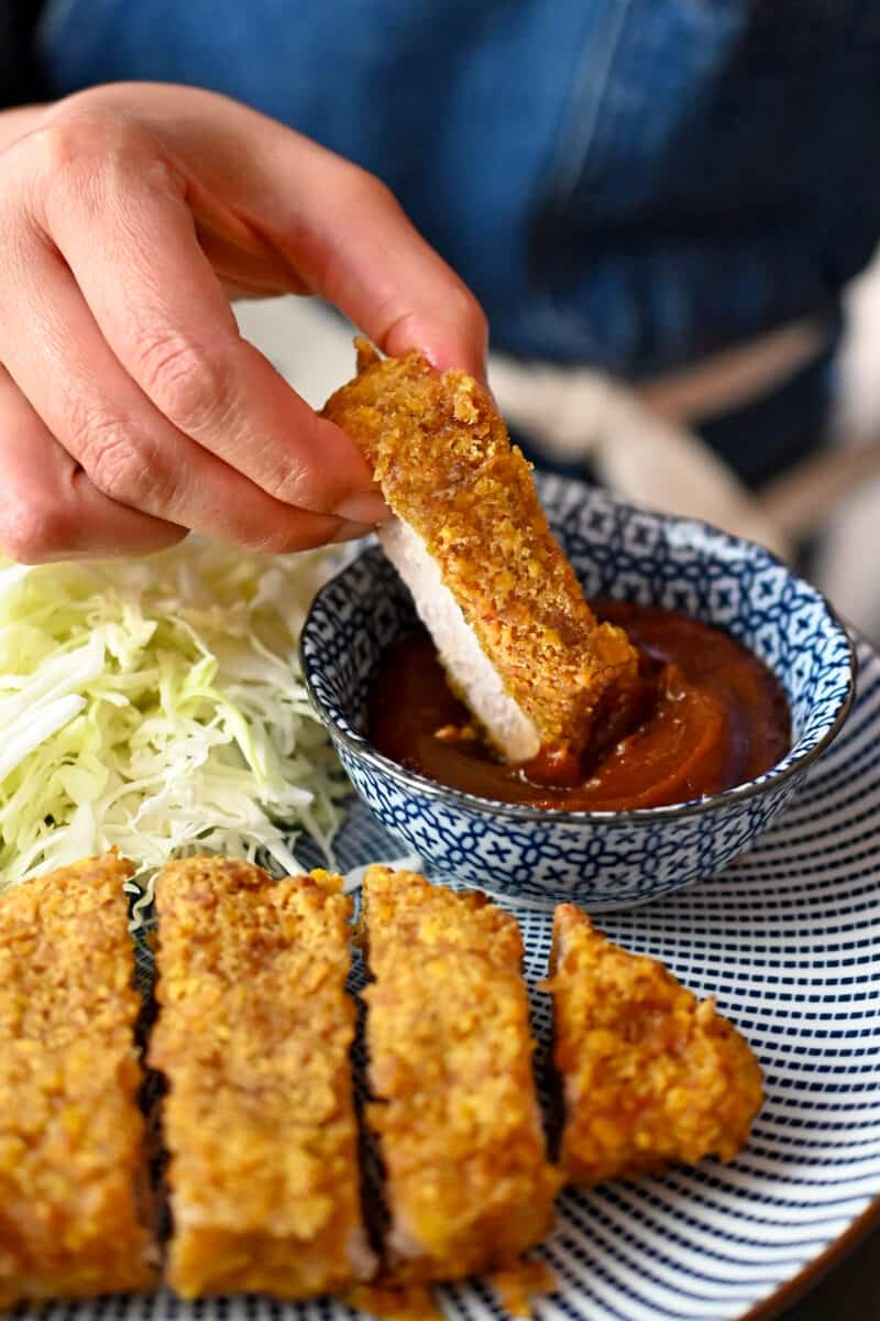 A hand is dipping a piece of pork katsu into a bowl filled with homemade Tonkatsu Sauce.