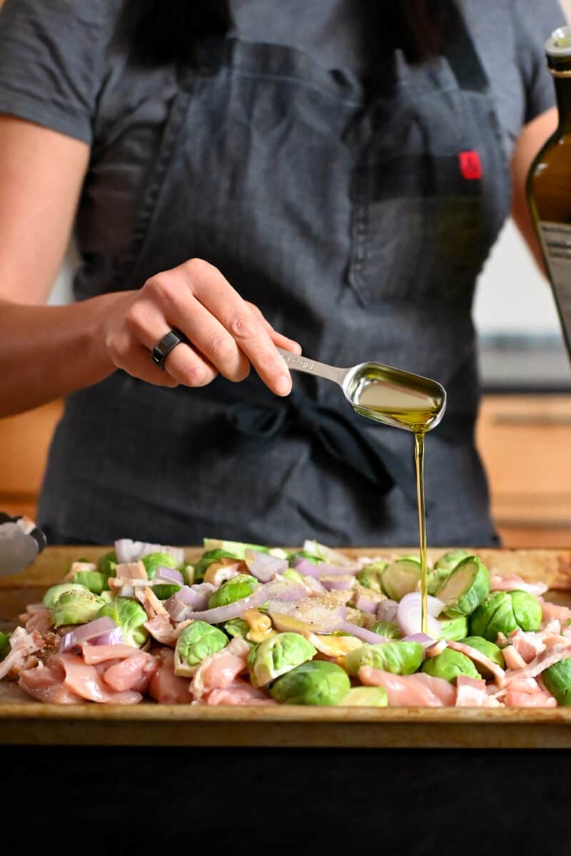 Adding a spoonful of extra virgin olive oil to a sheet pan filled with chicken and veggies .