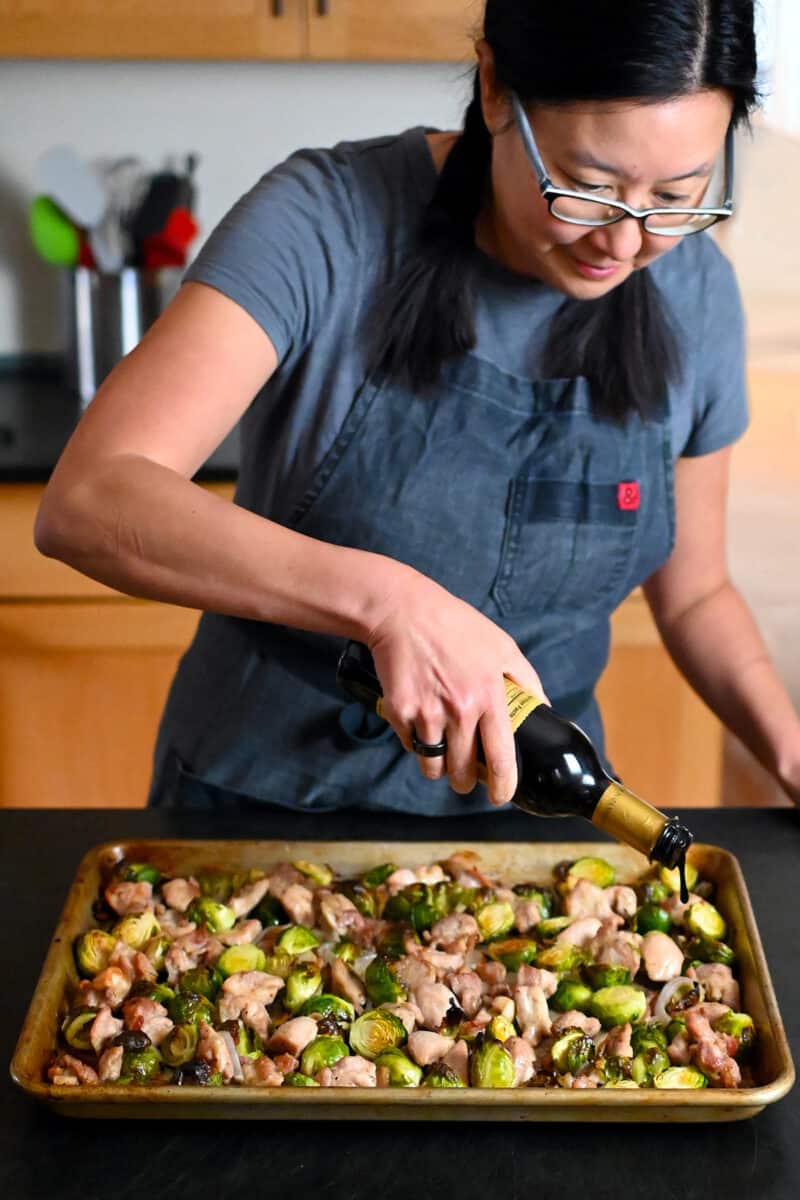 An Asian woman is drizzling aged balsamic vinegar on a sheet pan filled with chicken and veggies.