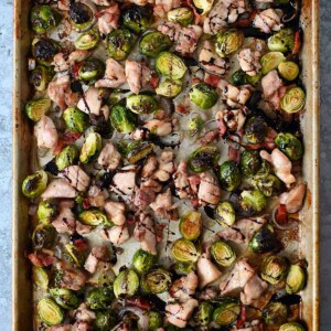 An overhead shot of a sheet pan with cubed chicken thighs, Brussels sprouts, bacon bits, and shallots.