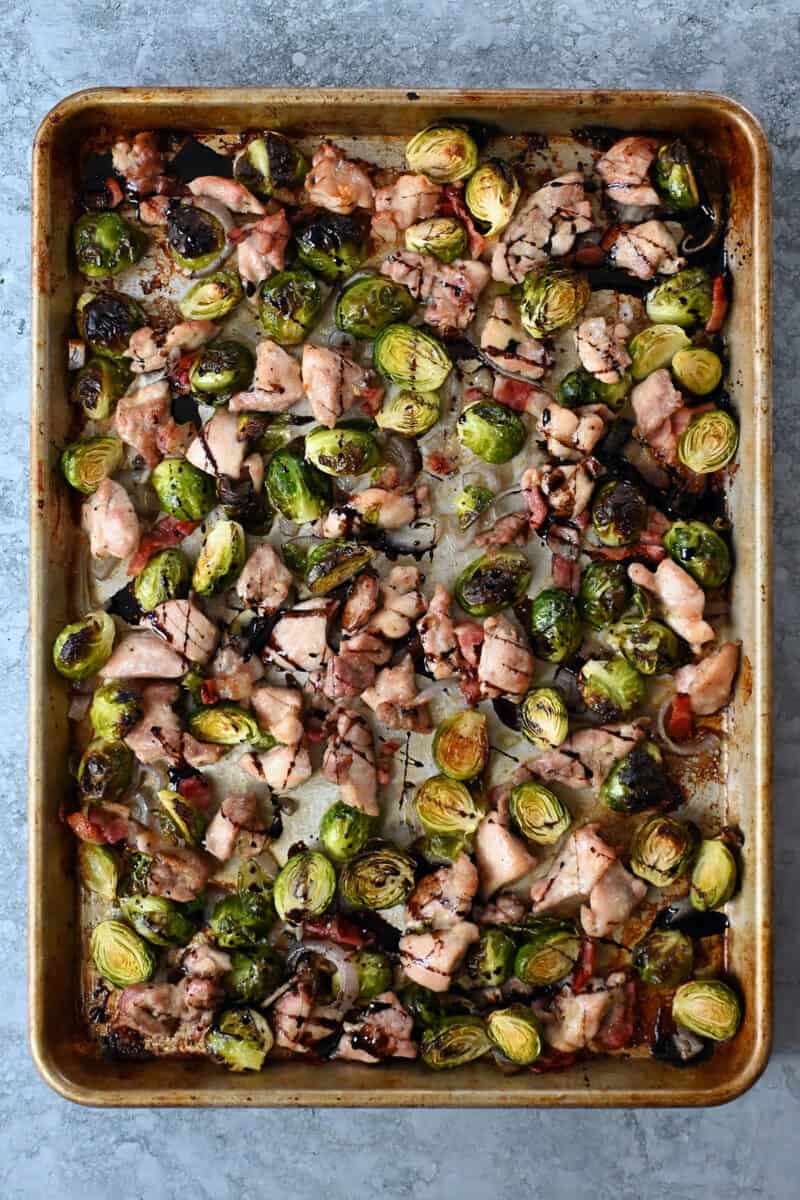 An overhead shot of a sheet pan with cubed chicken thighs, Brussels sprouts, bacon bits, and shallots.