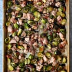 An overhead shot of two hands holding a sheet pan filled with chicken, Brussels sprouts, and bacon.