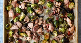 An overhead shot of two hands holding a sheet pan filled with chicken, Brussels sprouts, and bacon.
