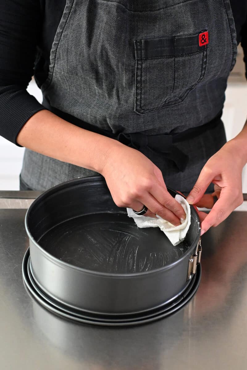 A person in a gray apron is greasing a springform pan with coconut oil.