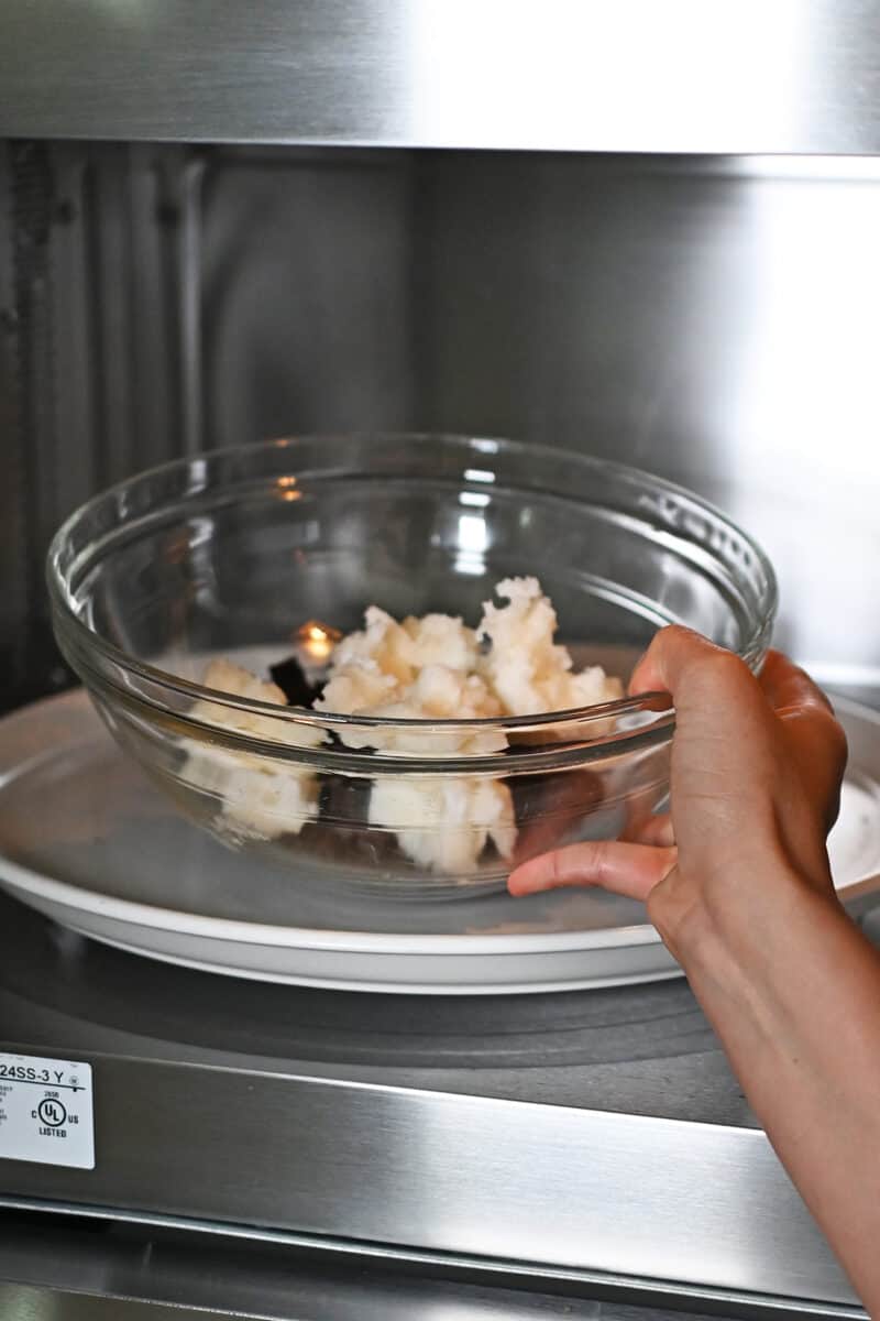 A hand is placing a large glass bowl filled with chopped dark chocolate and coconut oil into a microwave oven.