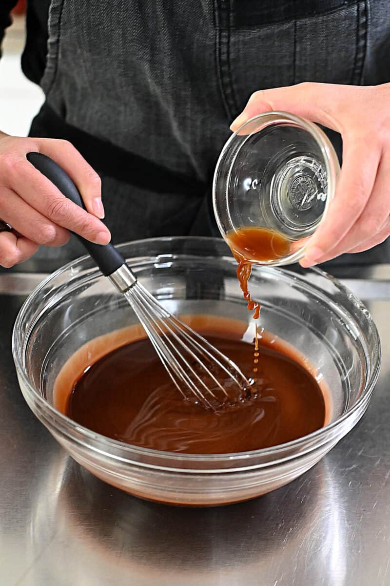 Pouring in a teaspoon of vanilla extract into a bowl of melted chocolate and coconut oil.