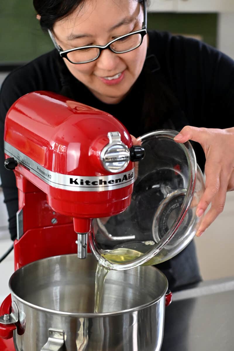 A smiling Asian woman in glasses is pouring egg whites into the mixing bowl of a red stand mixer.