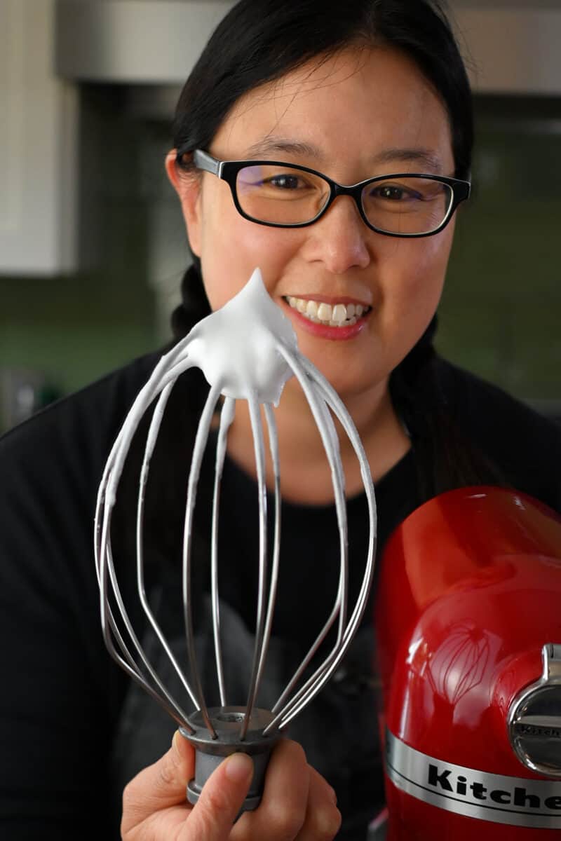 An Asian woman is holding up a whisk that has some whipped egg whites at the tip that are stiff and hold its shape.