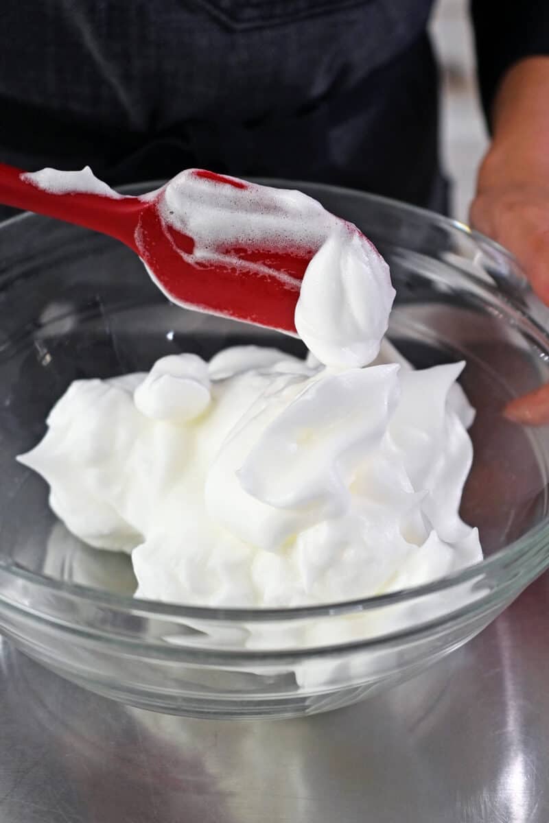 A red silicone spatula is transferring whipped egg whites to a large bowl.