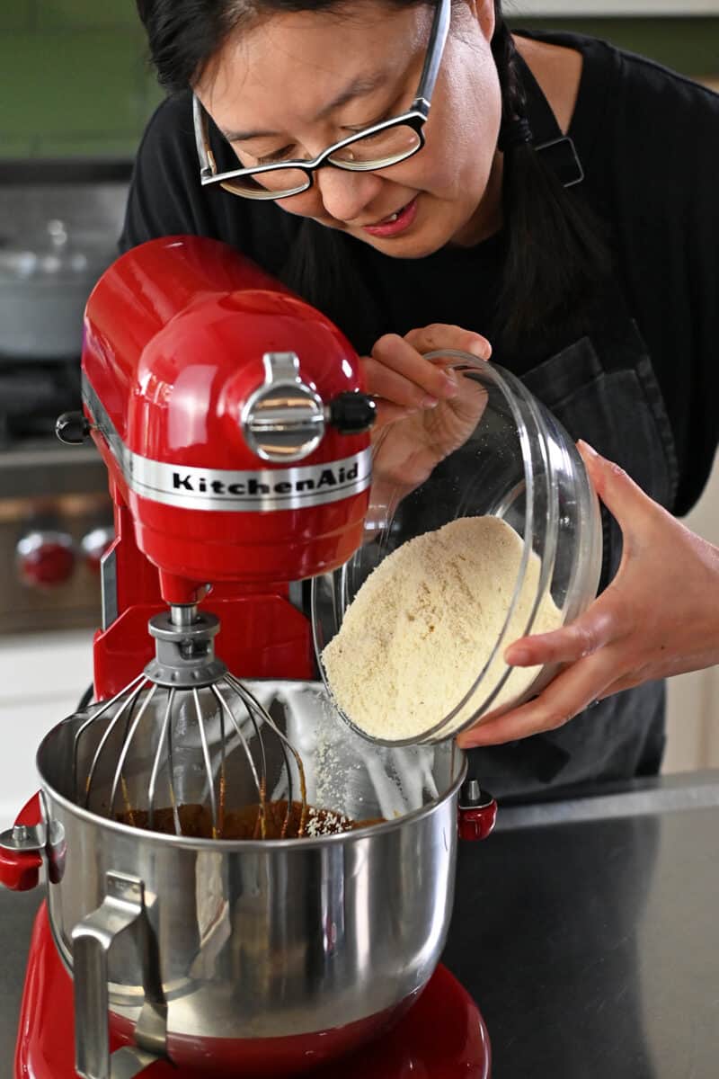 An Asian woman is adding almond flour to a stand mixer filled with Torta Caprese batter.
