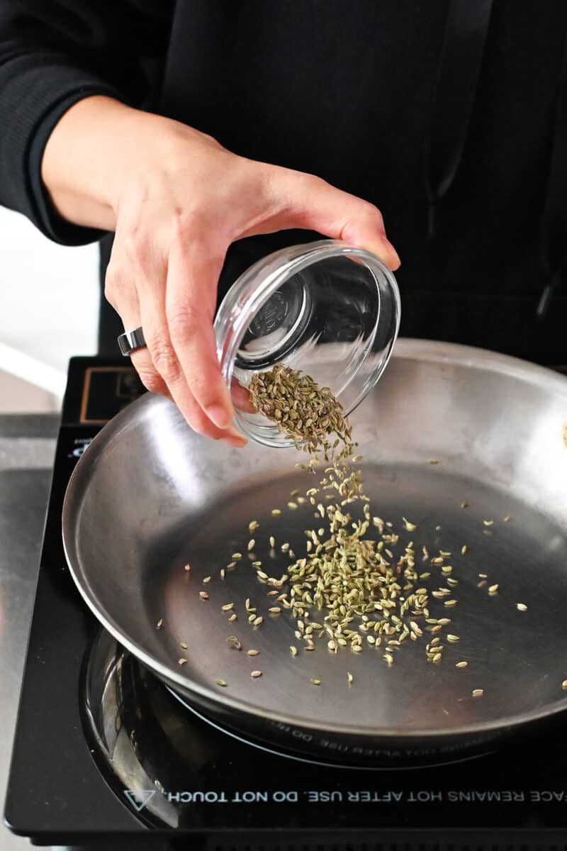 A hand is pouring fennel seeds into a stainless steel skillet.
