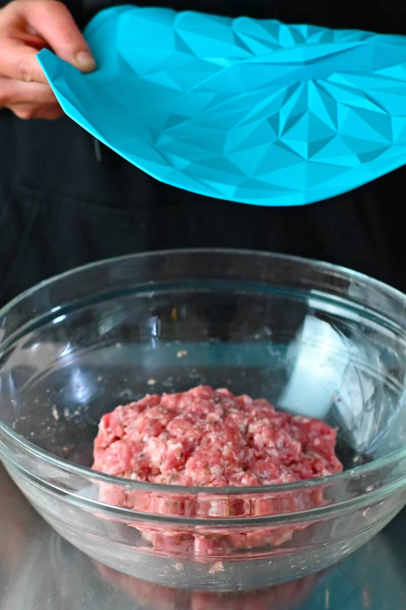 Two hands are placing a blue silicone lid on a bowl of raw Italian sausage that will be stored in the fridge.