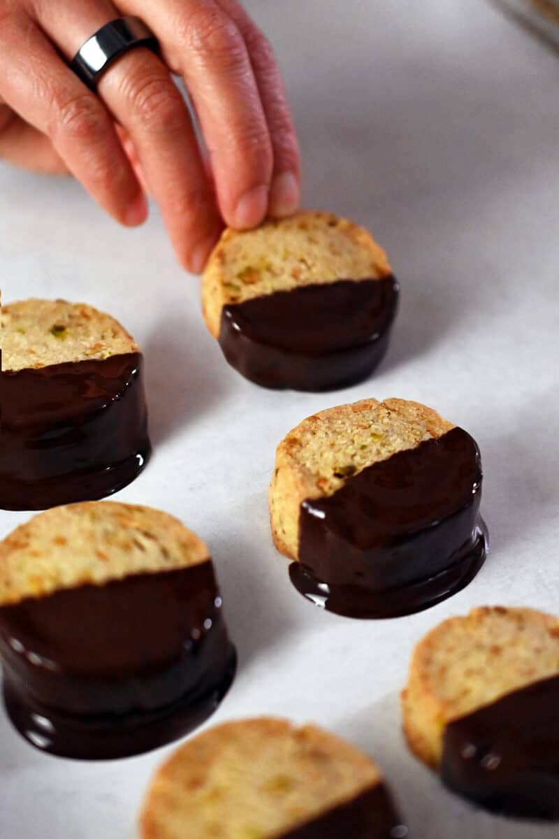 A hand is placing a chocolate dipped cookie onto a parchment lined baking sheet.