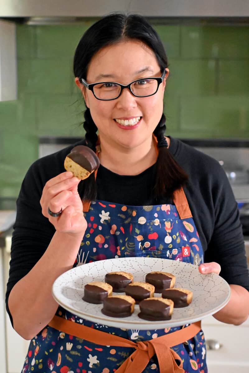 A smiling Asian woman holding a plate of chocolate dipped pistachio cookies. 