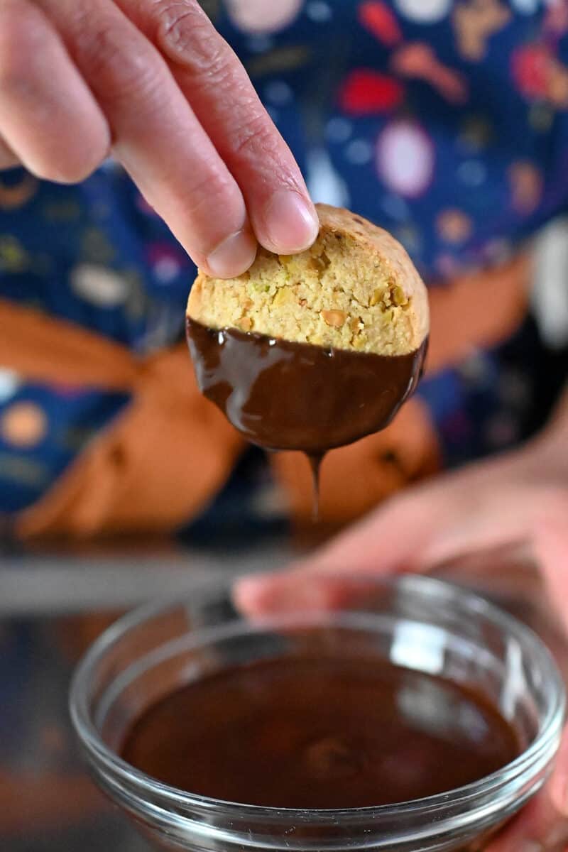 A hand is dunking a pistachio cookie into a bowl filled with melted chocolate.