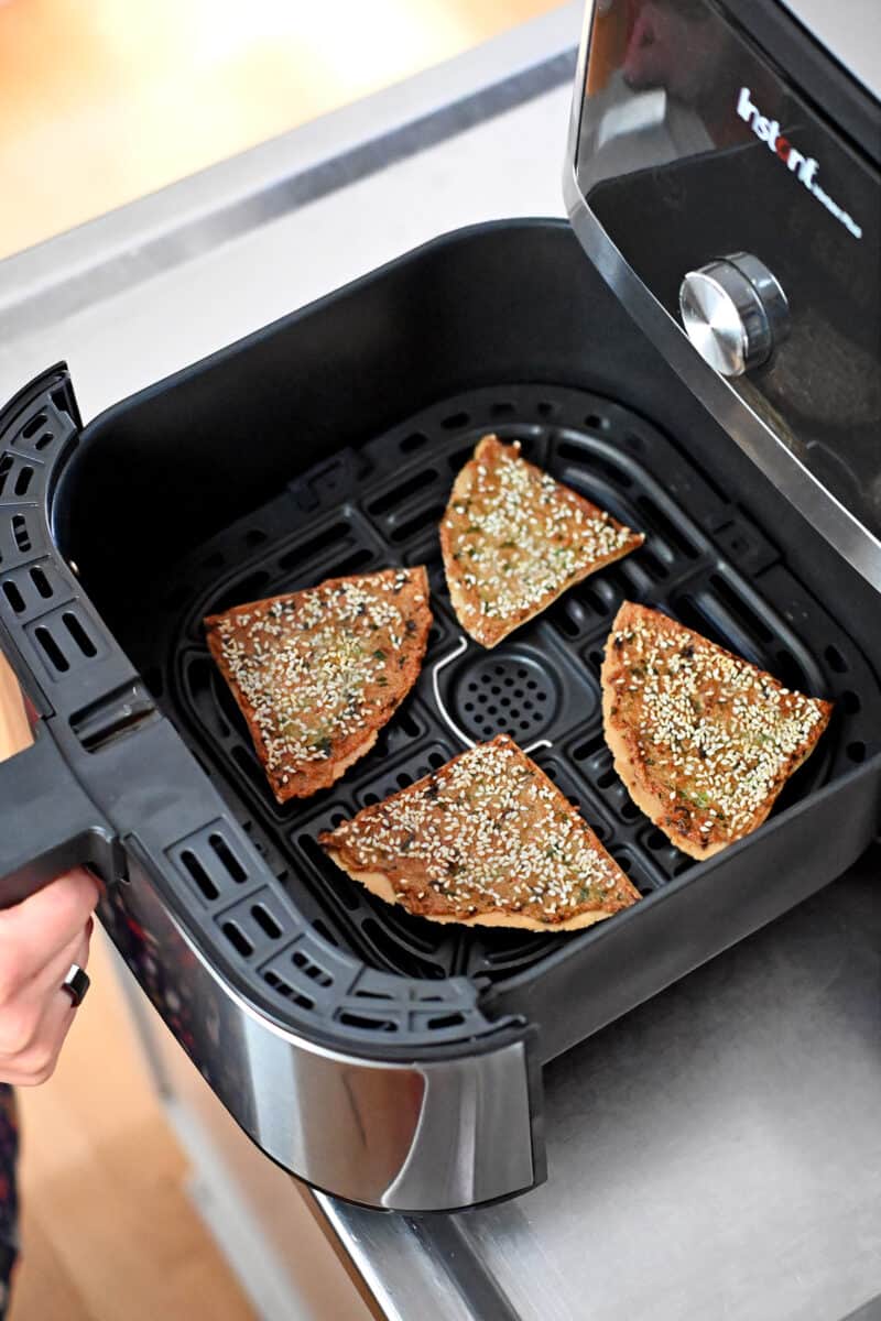 A hand is holding an air fryer open with four golden brown wedges of paleo shrimp toast inside the basket.