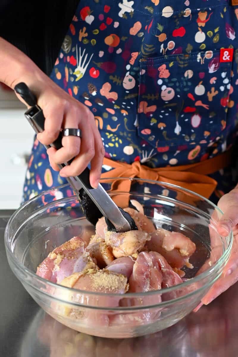 A person in a blue apron is tossing raw seasoned chicken thighs with a pair of tongs.
