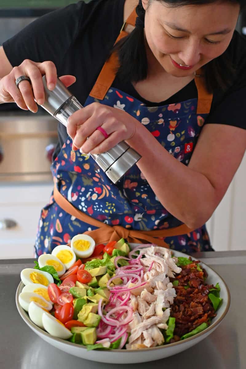 An Asian woman is grinding fresh black pepper on top of a Green Goddess Cobb Salad,