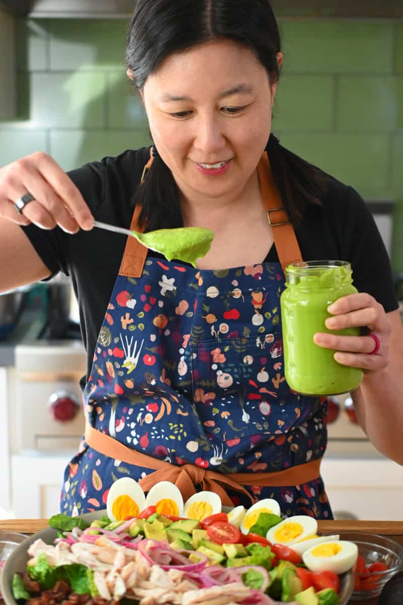An Asian woman in a blue apron is spooning Green Goddess dressing on a homemade Cobb Salad.