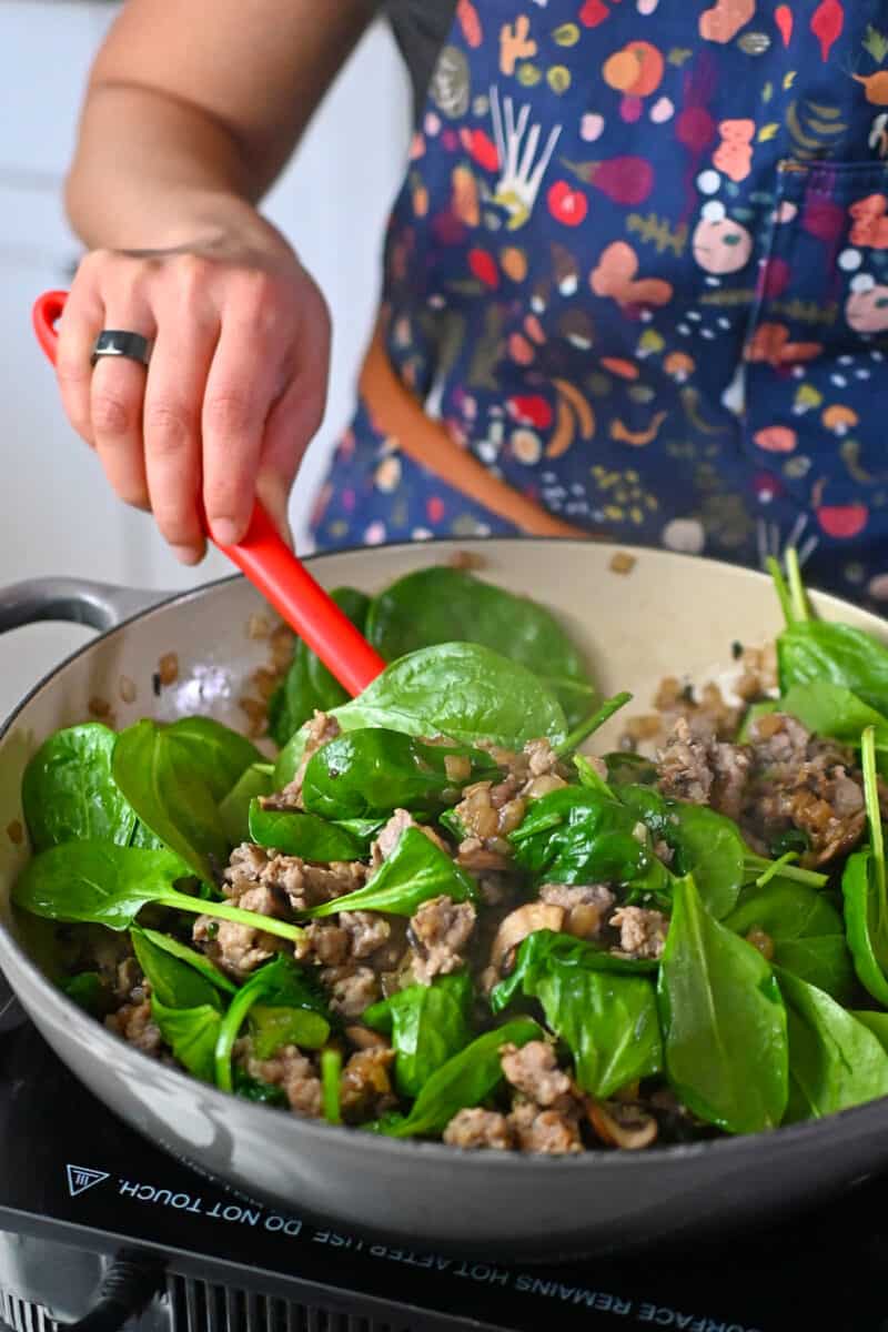 Stirring spinach broccoli into a skillet filled with cooked mushrooms, onions, and Italian sausage.