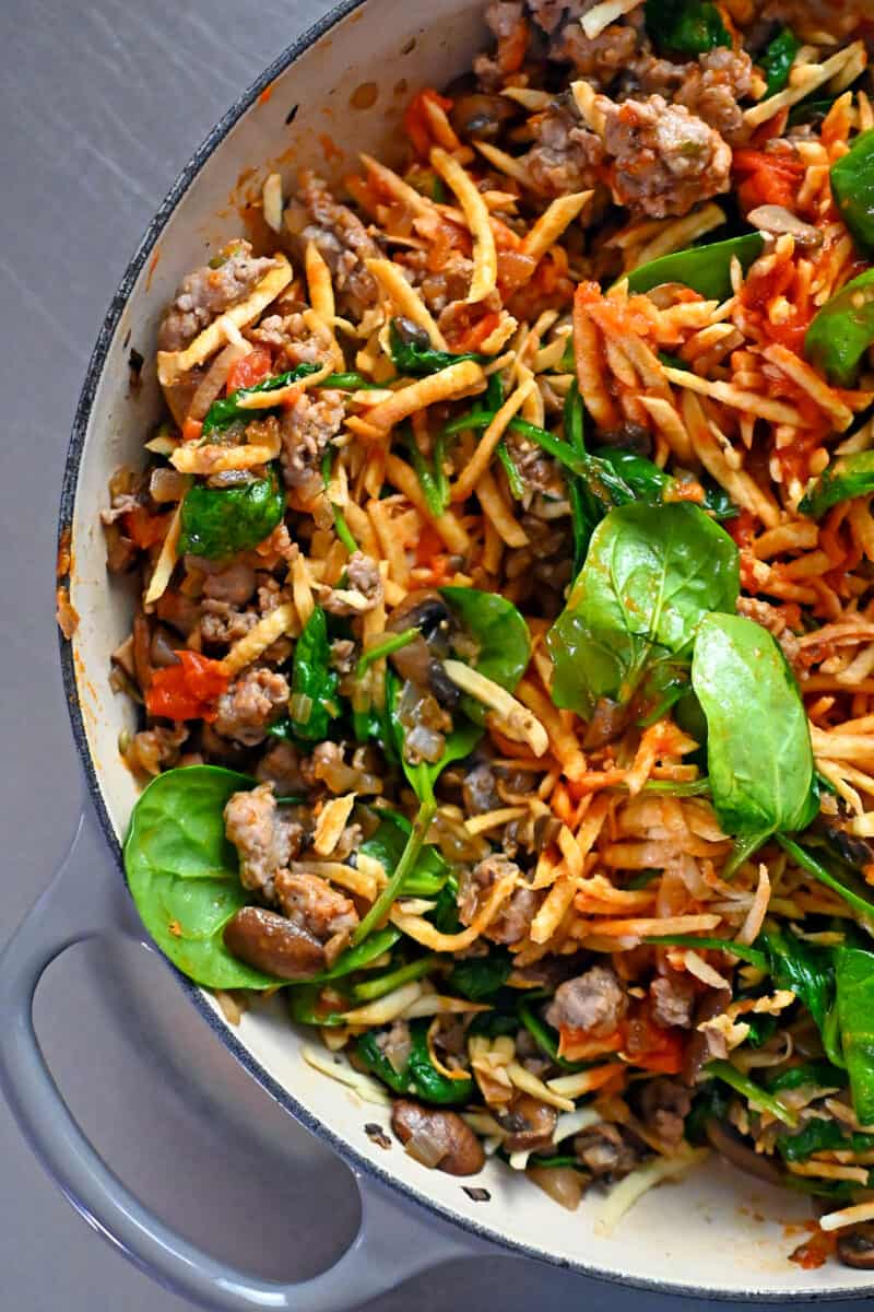 A skillet filled with Italian sausage, shredded white sweet potato, mushrooms, onions, and baby spinach.