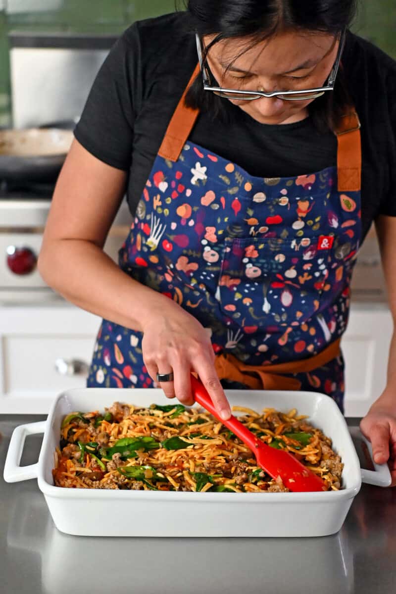 A woman in a blue apron is arranging cooked vegetables and sausage in a large baking pan.