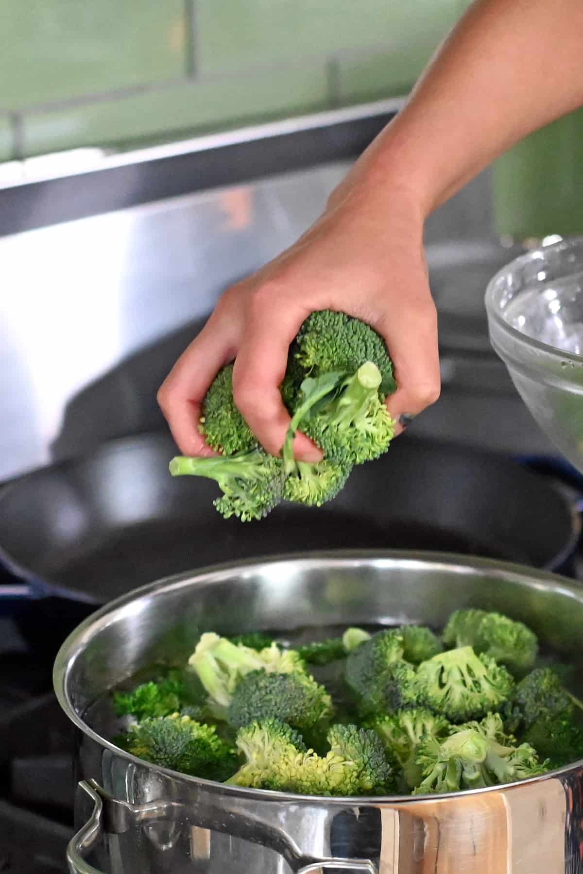 A hand is adding broccoli florets to an open pot of boiling water.