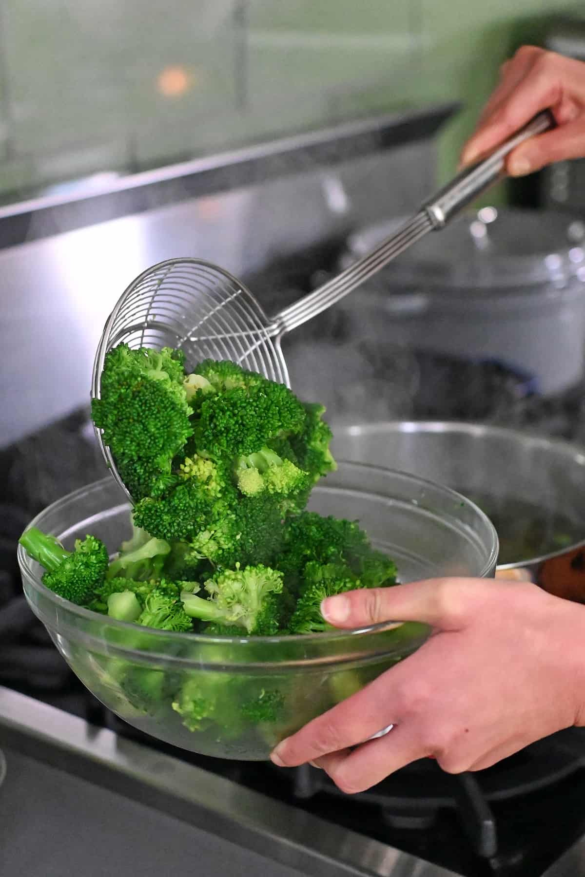 A metal sieve is being used to transfer cooked broccoli from a pot of boiling water to a clear class bowl.