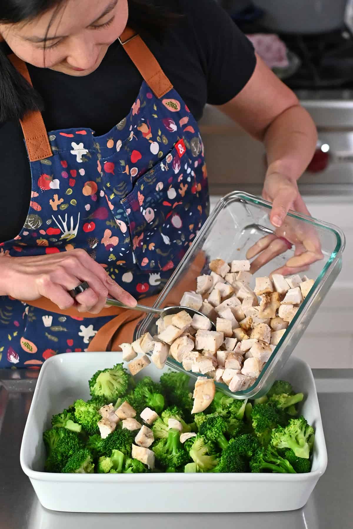 An Asian woman in a blue apron is adding cooked cubed chicken to a casserole dish with cooked broccoli florets. 