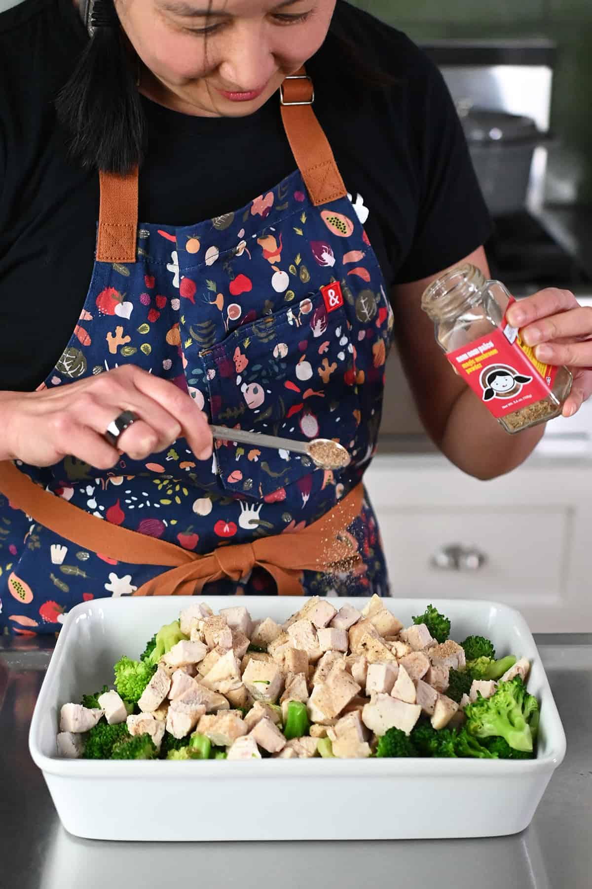 An Asian woman is adding some Nom Nom Paleo's Magic Mushroom Powder to a casserole dish with cooked chicken cubes and broccoli.