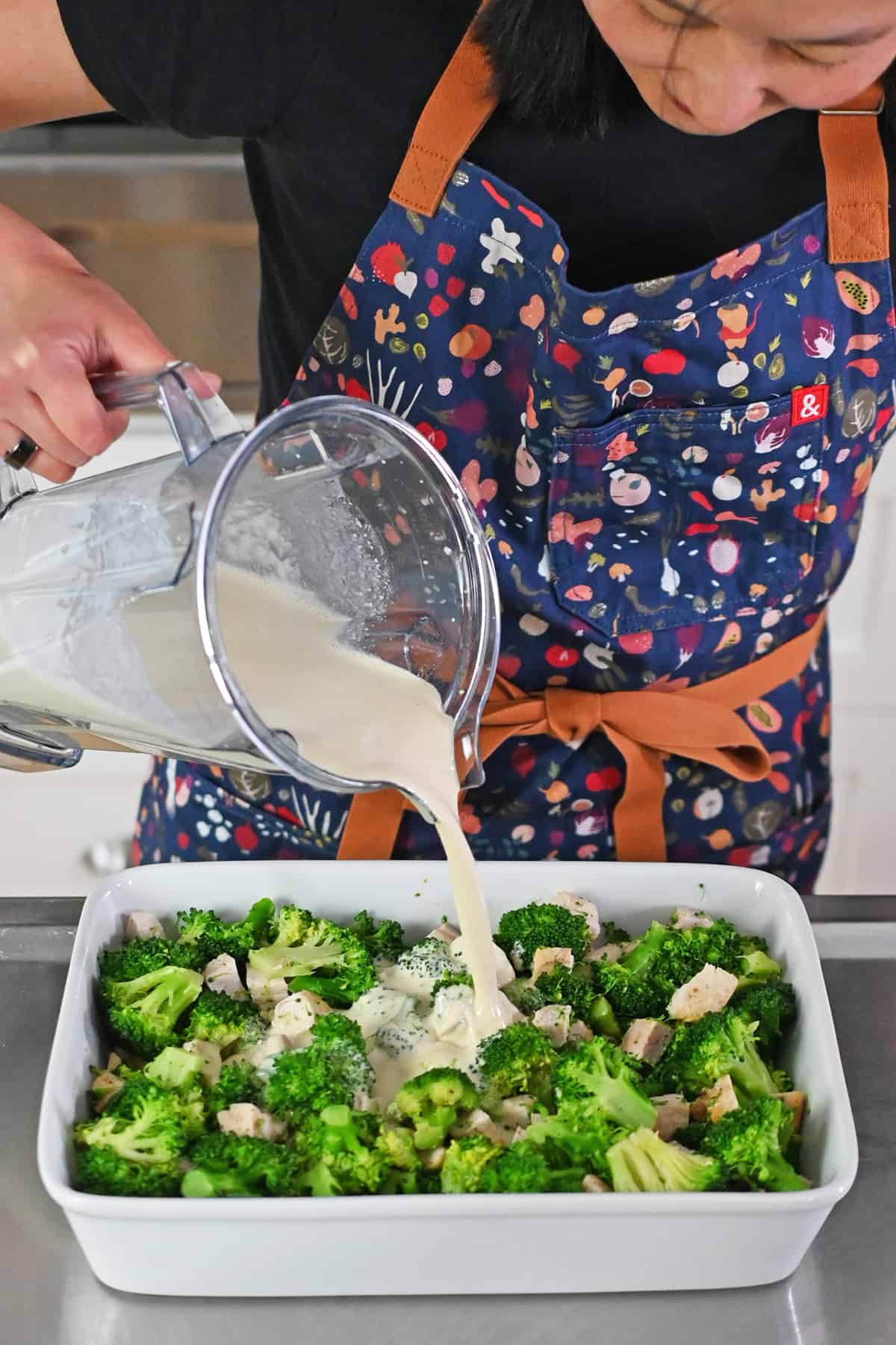 An Asian woman in a blue apron is pouring a white sauce from a blender onto broccoli and chicken in a white casserole pan.