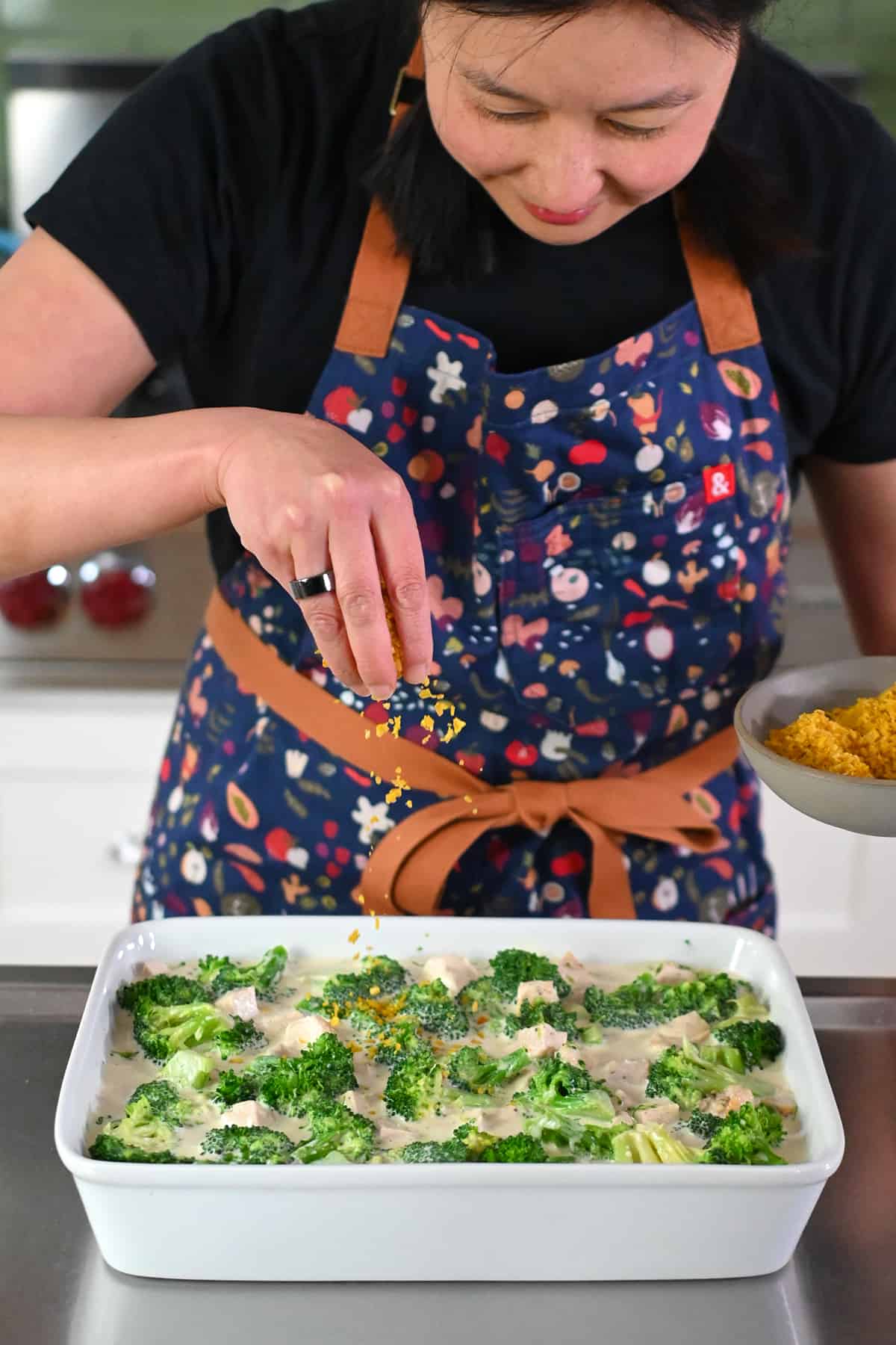 An Asian woman is sprinkling crushed plantain chips onto an unbaked paleo chicken divan casserole.