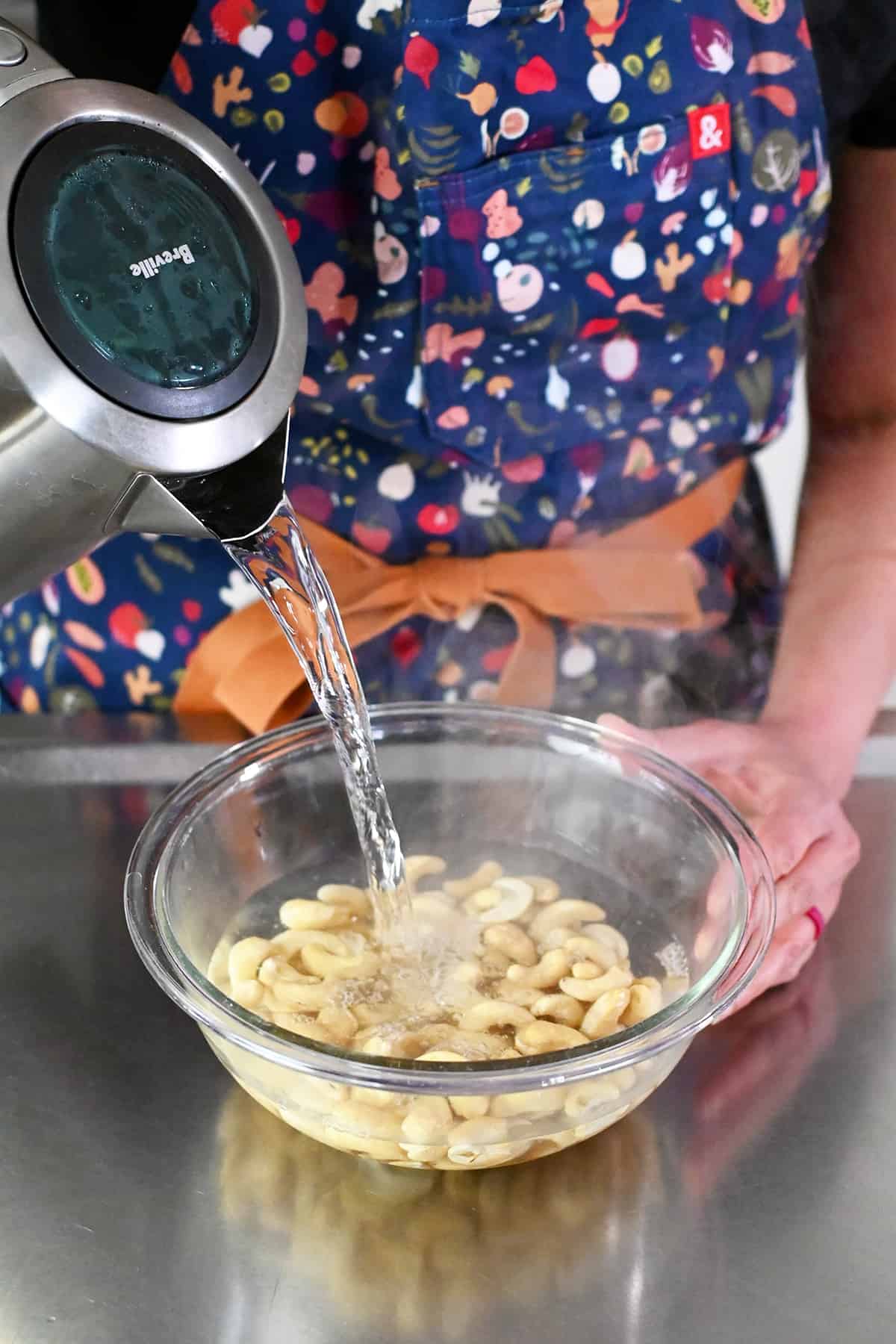 Pouring boiling water from an electric kettle into a clear glass bowl with raw cashews.