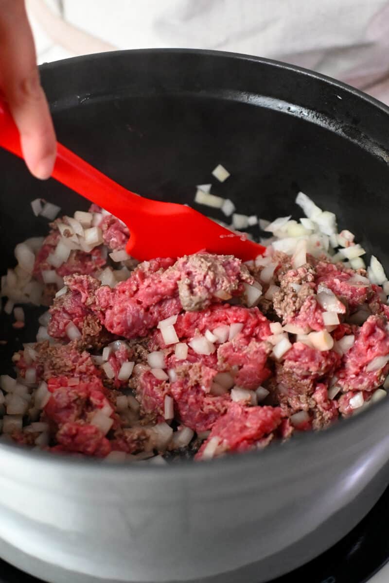 A red spatula is stirring some raw ground beef and diced onions in a large stockpot.