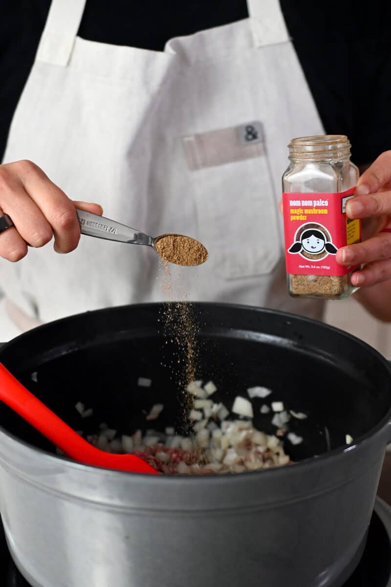 A person in a white apron is holding a jar of Nom Nom Paleo's Magic Mushroom Powder spice blend and adding half a teaspoon to a pot with cooked ground beef and diced onions.
