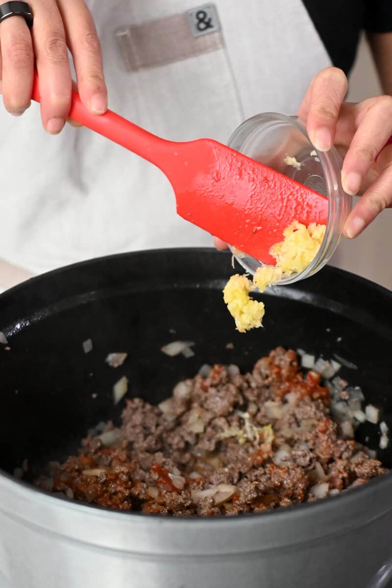 Adding minced garlic into a stovetop filled with cooked ground beef and diced onions.