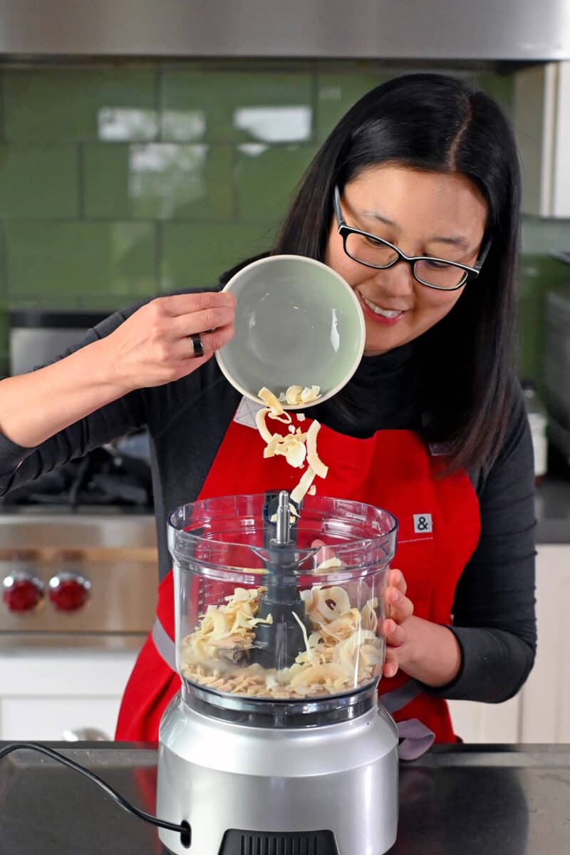 A smiling Asian woman is adding toasted coconut flakes into a food processor filled with toasted almond slivers.