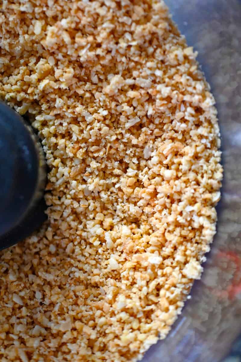 An overhead shot of an open food processor that shows the toasted almonds and coconut flakes are cut into a coarse cornmeal texture.