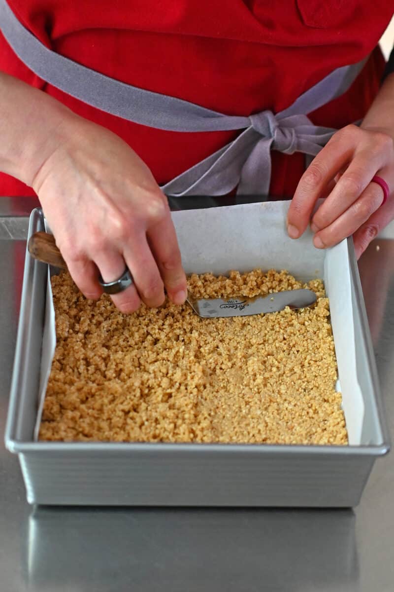 A hand is using an offset spatula to press a coconut and almond crust into the bottom of a parchment lined square baking sheet.